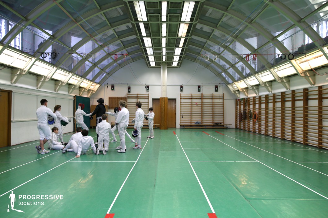 This compact fencing hall features a distinctive barrel-vaulted ceiling constructed from translucent panels supported by metal framing, allowing diffused natural light to complement the fluorescent fixtures suspended along the central spine. The space is lined with green flooring marked for fencing lanes, enhancing visibility and function. Wall-mounted wooden stall bars line both sides of the hall, adding a traditional sports facility character and providing functional exercise elements. The room is intimate and focused, ideal for close-up action shots or training sequences. Its vintage-sport aesthetic and symmetrical geometry make it suitable for both athletic and stylized visual productions.