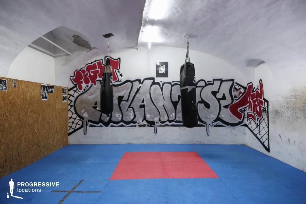 This gritty and compact underground boxing gym features a low vaulted ceiling and whitewashed walls that contrast with its raw, urban aesthetic. The space is fitted with two black punching bags suspended from ceiling mounts, placed above interlocking blue and red mat flooring that defines a central training zone. A large graffiti mural reading 