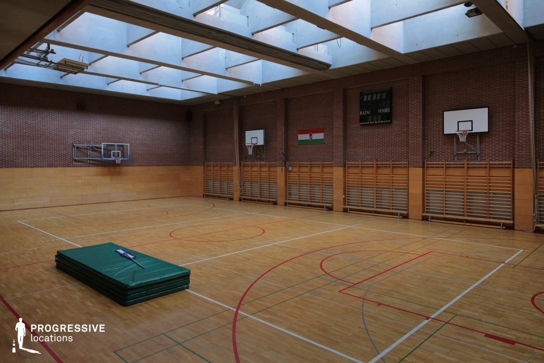 This image showcases a traditional indoor gymnasium featuring a polished wooden sports floor marked with colorful lines for multiple disciplines, including basketball and volleyball. The space is enclosed by high brick walls with a horizontal wooden panel section at the lower half, creating a warm and structured aesthetic. Mounted basketball hoops are evenly distributed across the walls, and a mechanical scoreboard is positioned prominently above one side, reinforcing the venue's formal athletic purpose. Overhead, a series of translucent ceiling panels in rectangular recesses allows diffused natural light to flood the space, giving it a balanced and well-lit atmosphere. Exposed beams and modern structural elements contribute to a functional and minimalistic design. Wall-mounted wooden climbing frames line the gym's perimeter, adding to its versatility for various physical activities. The dimensions are generous, offering an unobstructed central playing field suitable for wide shots or choreographed movements. Its clean architectural lines and neutral tones make this location highly adaptable for sports-themed productions, physical training sequences, or institutional backdrops in film and photography.