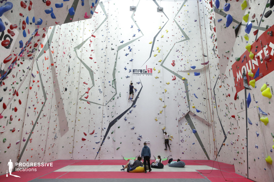 This expansive indoor climbing facility features towering climbing walls with varied overhangs, volumes, and textures, all outfitted with multicolored holds arranged across numerous routes. The floor is cushioned with vibrant red and gray crash mats, offering both visual structure and safety. The white and light-gray angular walls are punctuated by bold graphic lines and brand elements, enhancing the contemporary aesthetic. Ceiling-mounted auto-belays and top ropes support different climbing skill levels. The venue is ideal for sports shoots, dynamic action scenes, or lifestyle campaigns centered on athleticism and urban adventure.