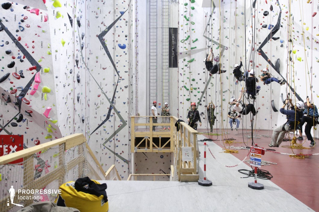 This image showcases a large indoor climbing facility designed for both sport and rope access training. The hall features towering white climbing walls outfitted with multicolored holds and overhang structures arranged in geometric patterns. The walls reach significant heights, maximizing the vertical scale and offering a visually dynamic backdrop. The textured routes are marked with vibrant colors including neon yellow, pink, green, and red, enhancing the location’s visual energy. The space is organized into climbing zones with a designated red rubber floor and suspended rope systems for vertical maneuvering and safety instruction. A wooden platform structure with railings creates a mid-level observation or rest area, adding depth and layering to the spatial composition. The ceiling height and wall continuity offer flexibility for wide framing and vertical camera movement. Combining bold design, clear functional zoning, and strong architectural rhythm, this venue is ideal for energetic and high-impact visual projects such as sports documentaries, action-oriented scenes, or performance-based advertising.