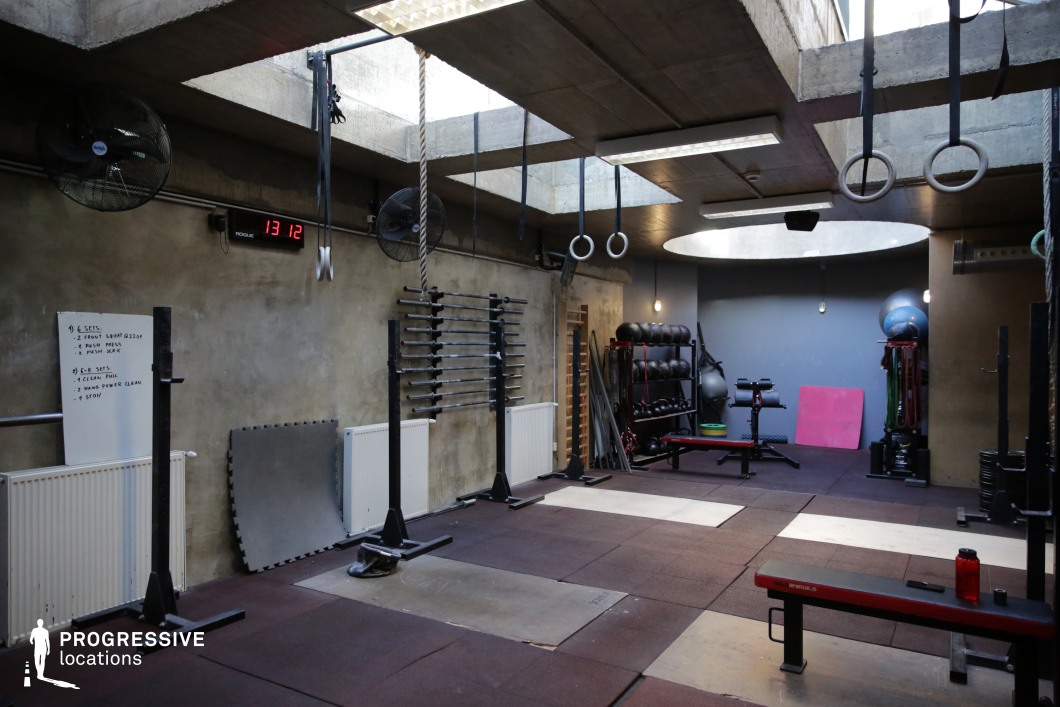 A subterranean-style gym space with raw concrete ceilings and skylights, this location combines industrial aesthetics with functionality. The room is outfitted with Olympic lifting platforms, barbell racks, wall-mounted equipment storage, gymnastics rings, and rope climbs. The distressed concrete walls and minimalistic lighting give the interior a gritty, performance-focused atmosphere. Padded rubber flooring covers the entire area, suitable for high-impact workouts and functional training routines. The open ceiling cut-outs allow natural light to stream in, creating dramatic contrast and enhancing the visual depth. This venue is ideal for scenes centered around intense athleticism, underground sports culture, or personal transformation.