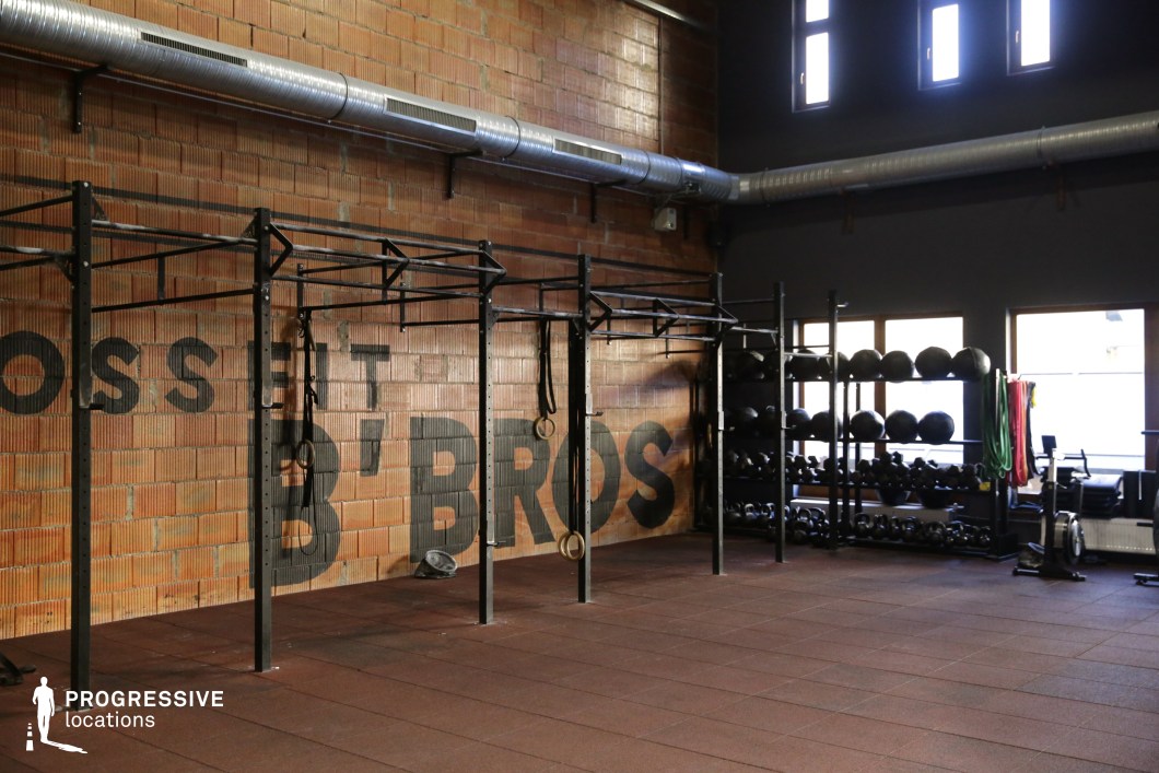 This CrossFit-style gym features an industrial look defined by its exposed brick walls, high ceilings, and visible ventilation ducts. The space is equipped with a full wall-mounted pull-up and rig structure, wall balls, kettlebells, and resistance bands neatly organized on open steel racks. Large windows provide ample natural light, while the combination of matte flooring tiles and dark-painted upper walls contributes to a modern yet raw atmosphere. The space is open-plan and versatile, suitable for high-intensity workouts, fitness-focused shoots, or scenes depicting urban athletic training. The minimalist, rugged setting supports dynamic camera movement and functional staging.