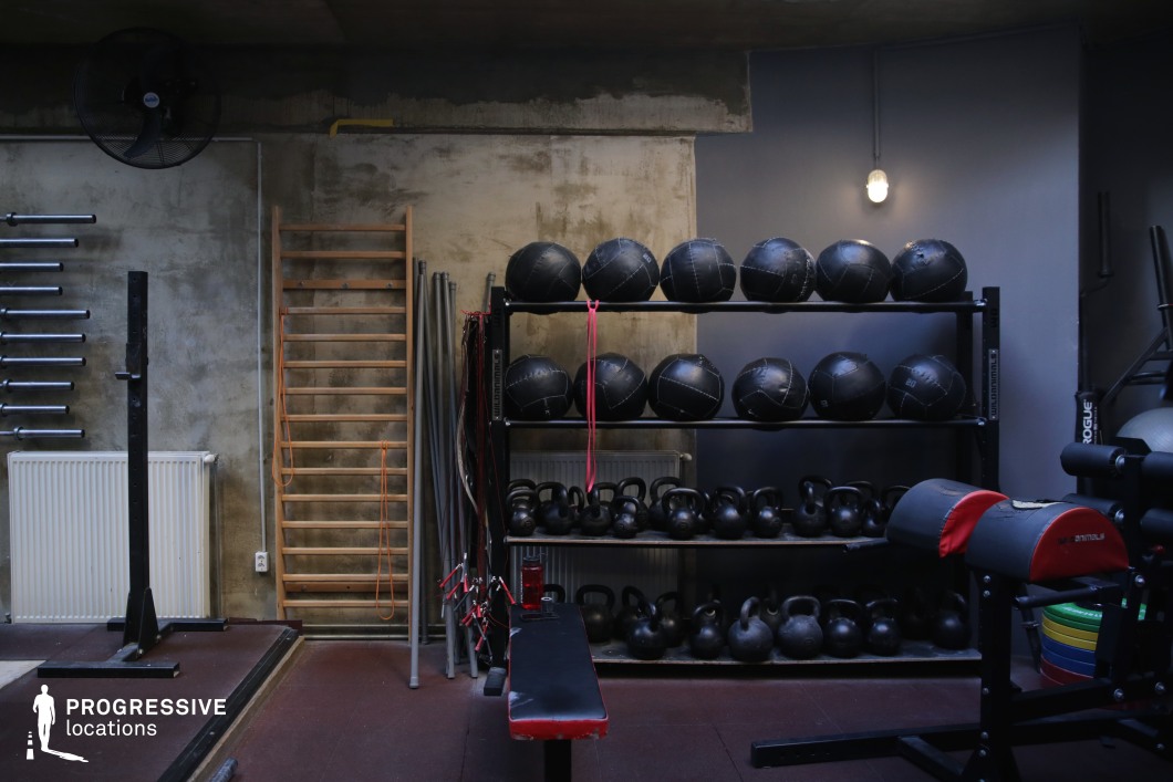 This compact, industrial-themed training area features a rugged interior with exposed concrete walls, dark flooring, and focused ambient lighting. The space is equipped with a full rack of medicine balls, kettlebells, and weightlifting bars, organized neatly for functional fitness routines. A red-accented Roman chair and flat bench contribute to the room’s utilitarian feel. Textured wall elements, a mounted wooden stall bar, and a subtle mix of metallic and wooden finishes enhance the raw atmosphere. Ideal for scenes requiring an underground gym aesthetic or character-driven athletic montages, this location offers a gritty yet structured visual tone.