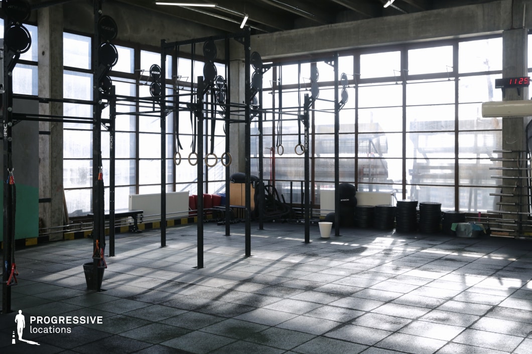 This open-plan, loft-style training facility combines industrial architecture with contemporary fitness design. It features high concrete ceilings, expansive floor-to-ceiling windows, and durable black rubber flooring throughout. The central rig structure supports gymnastic rings, pull-up bars, and various suspension training setups, complemented by neatly stacked weight plates, medicine balls, and resistance ropes in the background. The space is illuminated by sleek linear LED fixtures and flooded with daylight, making it visually dynamic and production-friendly. The minimalist, raw aesthetic and functional layout make it a versatile filming location for athletic brand campaigns, high-energy workouts, or urban fitness montages.