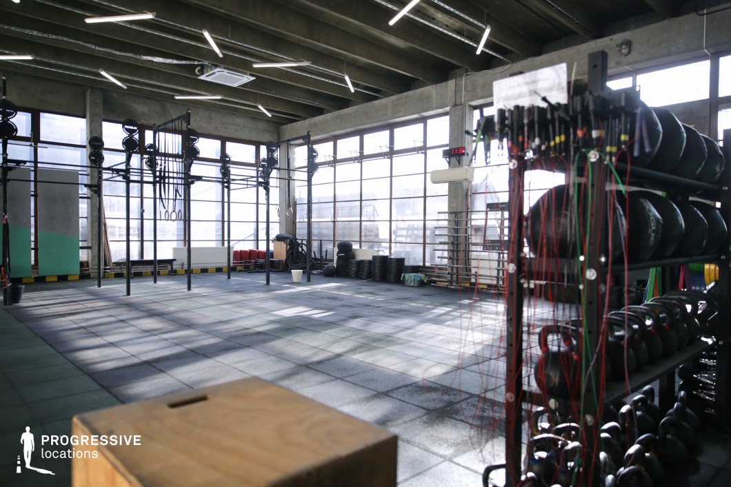 This spacious, industrial-style CrossFit gym features an open layout with high ceilings, exposed concrete beams, and floor-to-ceiling windows that allow natural light to flood the space. The flooring consists of heavy-duty black rubber tiles suitable for high-impact training. The gym is outfitted with modular rig structures, pull-up bars, wall balls, resistance bands, kettlebells, and plyometric boxes, offering a fully equipped environment for functional fitness. The raw concrete surfaces, linear lighting fixtures, and minimalist aesthetic contribute to a modern urban atmosphere, making it an ideal location for dynamic sports commercials, fitness brand shoots, or contemporary training montages.