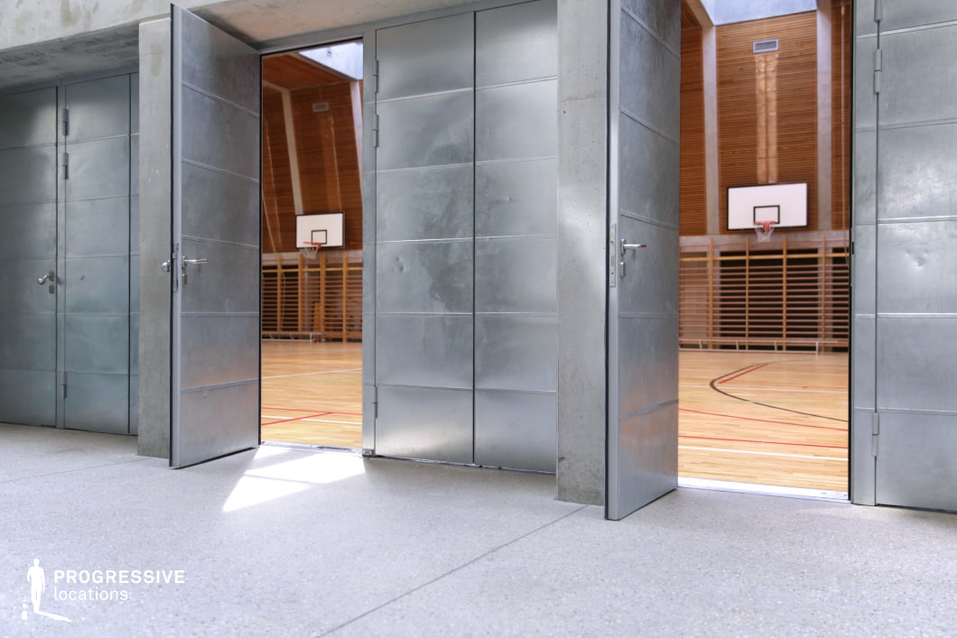 This image captures a unique transitional space between a concrete corridor and a contemporary gymnasium, framed by large industrial-style metal doors. The open doors reveal the interior of the gym hall, which is clad in warm-toned wood paneling that extends vertically across the high walls, establishing a striking contrast with the raw, cool textures of the concrete and steel foreground. Inside, the gymnasium floor is finished in polished light wood with red and black court markings, and standard basketball hoops are mounted against evenly spaced vertical slats. The space feels both robust and refined, balancing hard architectural elements with natural materials. The symmetrical composition, juxtaposition of textures, and the play of light between interior and exterior make this location particularly suited for cinematic entries, architectural photography, or conceptual visual storytelling focused on contrast and transition. Its minimal yet bold structure offers high adaptability for creative productions.