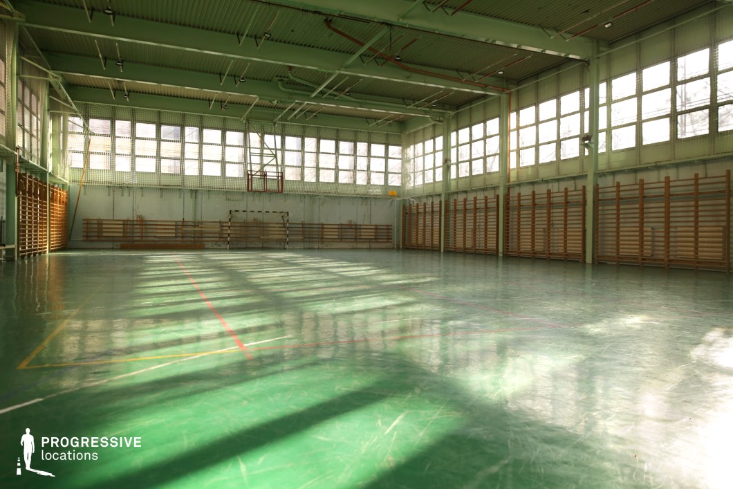 This image shows a spacious indoor gymnasium with a distinctly retro aesthetic, characterized by its polished green sports flooring and high, industrial-style ceiling. The walls are lined with large rectangular windows on two sides, allowing ample natural light to stream in and cast geometric shadows across the floor. Wooden wall bars line the perimeter, hinting at a school or recreational setting. The space is framed by mint green-painted steel beams and a metal-paneled ceiling with exposed piping, enhancing its utilitarian charm. The court markings and central goal indicate multifunctional use, ideal for basketball, handball, or indoor physical education. Its clean, nostalgic design and abundant light make it a fitting backdrop for scenes requiring a classic, institutional athletic environment.