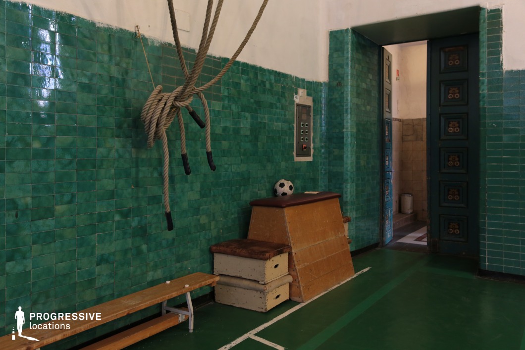 This image captures a detailed corner of a vintage gymnasium interior, showcasing distinctive green-glazed ceramic wall tiles and original gym equipment, such as a wooden vaulting box, gym ropes, and a classic bench. The entrance features elaborately paneled doors and a partially visible tiled corridor, enhancing the setting's historical character. The worn textures, muted lighting, and presence of a small control panel embedded in the wall suggest minimal modernization, preserving the location’s retro authenticity. Ideal for period film scenes, nostalgic sports settings, or stylized commercial shoots requiring a distinctive old-school gym atmosphere.