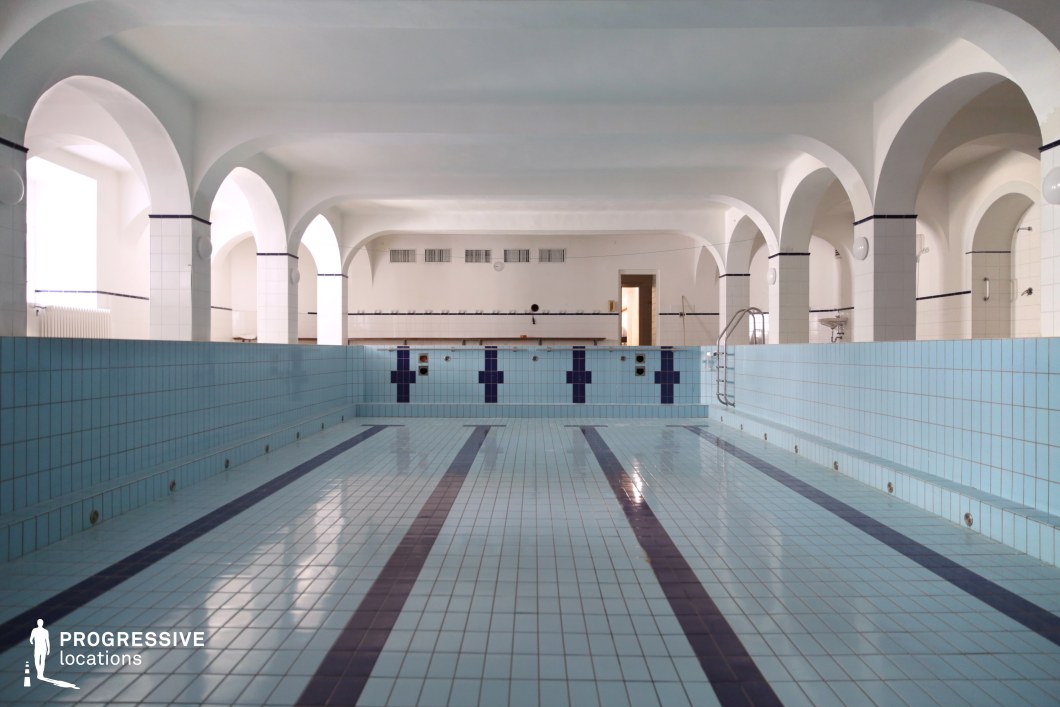 This image presents a distinctive indoor swimming pool space characterized by its classic architectural elements and serene atmosphere. The pool is currently drained, revealing a tiled interior composed of pale blue surfaces accented with deep blue lane markers and vertical design elements. The layout is symmetrical, guiding the viewer’s eye toward the far end, where a central doorway and evenly spaced ventilation units form a natural visual anchor. A defining feature of the space is the series of white, rounded arches supported by square columns that frame the pool area. These structural elements add rhythm and a sense of historical depth to the room. The walls and ceiling are finished in a clean, matte white, amplifying the light and enhancing the spatial clarity. The tiled surfaces and built-in steps emphasize the facility’s functionality, while the balanced proportions and geometric precision offer a visually calm and formal aesthetic. This location is ideal for production needs requiring a refined, enclosed aquatic setting with timeless architectural character and clean visual lines.