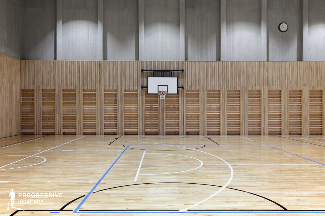 This contemporary gymnasium presents a clean, minimalistic interior with light-toned wooden flooring and slatted wood wall paneling, creating a visually cohesive and warm atmosphere. The court is marked with black, blue, and white lines, supporting multi-sport use such as basketball and volleyball. A centrally placed basketball hoop is mounted against the wood-paneled wall, flanked by a series of integrated wooden stall bars that enhance the functional design. The upper wall section transitions to exposed concrete with vertical insets, adding architectural rhythm and industrial refinement. A single analog clock on the far wall offers a practical focal point. This versatile and acoustically considered space is ideal for dynamic sports-themed shoots or as a neutral, modern background for commercials and visual media.