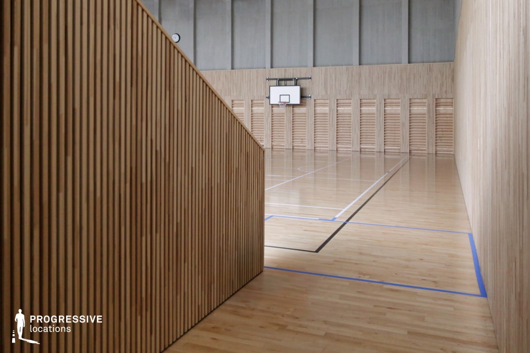 This location showcases a modern indoor sports hall defined by its minimalistic and natural design elements. The walls and floors are clad entirely in light-toned wood, creating a unified and calming visual identity. Vertical slatted wooden panels run along the walls and staircase, offering texture and depth while maintaining clean lines throughout the space. The flooring is a polished hardwood surface marked with subtle blue and black game lines, enhancing both functionality and aesthetic clarity. A single basketball hoop is mounted against a backdrop of continuous wooden slats, emphasizing symmetry and focus within the composition. The tall ceilings and neutral grey upper walls give the space an airy, acoustically balanced atmosphere. The restrained palette and strong geometric structure provide an ideal backdrop for creative projects requiring a sleek, Scandinavian-inspired athletic interior. This setting is particularly suited for high-end fashion editorials, contemporary sports visuals, or architecturally focused film productions.