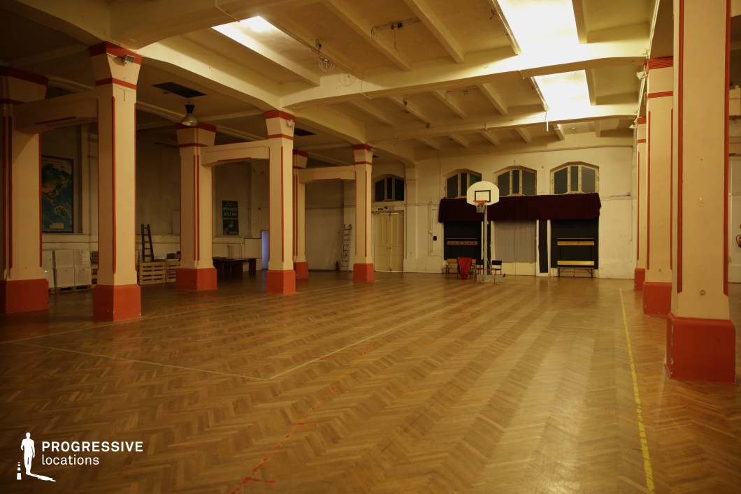 This atmospheric gymnasium features a parquet herringbone-pattern wooden floor paired with robust square columns painted in a pale cream tone with red accents. The space exudes an old-school charm, enhanced by its high ceilings with exposed beams and soft, warm lighting. Large arched windows, partially draped and framed by dark curtains, add character to the venue. A single basketball hoop stands at one end, underscoring the multifunctional use of the hall. The architectural style and slightly aged interior suggest historical significance, making it suitable for retro-inspired film scenes, nostalgic sports sequences, or artistic fashion productions in a timeless, versatile indoor environment.