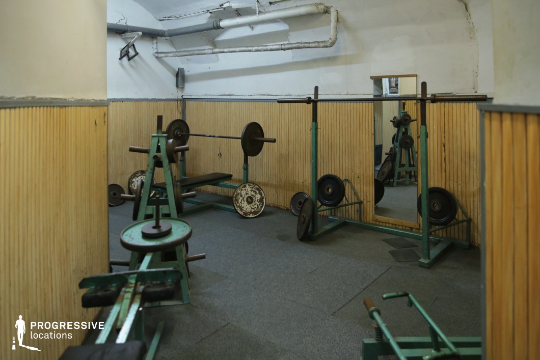 This compact gym area showcases a vintage weightlifting setup with teal-painted metal equipment and aged weight plates. The space features arched ceilings, exposed pipes, and vertically paneled wooden walls that create a utilitarian, basement-like atmosphere. Mirrors on the wall visually expand the room and enhance the raw, old-school ambiance. The muted tones, industrial textures, and simple flooring contribute to an authentic, gritty character, making it an excellent backdrop for underground training sequences, retro fitness content, or documentary-style productions emphasizing authenticity and discipline.