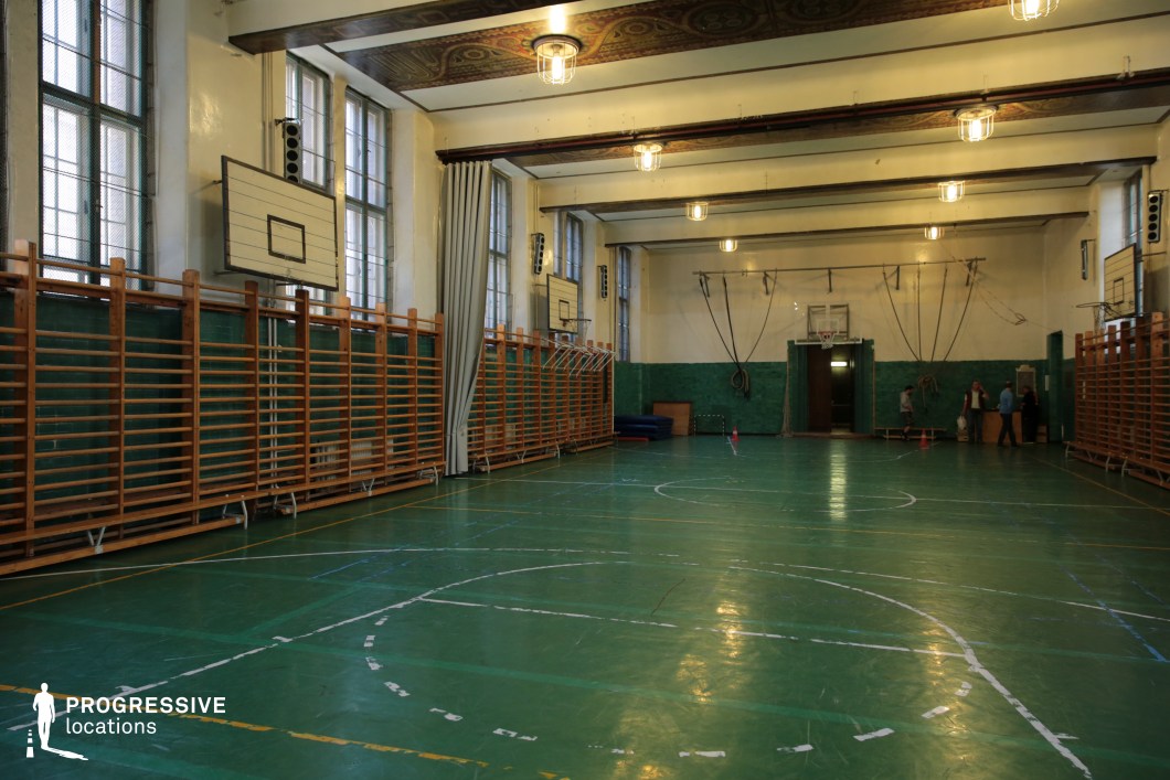 This vintage indoor gymnasium features high ceilings, detailed ceiling decorations, and large multi-pane windows that flood the space with natural light. The polished green sports flooring is marked for multiple sports, while traditional wooden wall bars line both sides of the hall, providing a classic athletic aesthetic. The gym’s aged charm is enhanced by mid-century basketball backboards and ceiling-mounted gymnastics equipment, such as hanging rings and ropes. The space is equipped with folding partitions and original architectural elements like decorative ceiling panels and framed beams. This gym is ideal for period productions, nostalgic sports settings, or scenes requiring an institutional yet character-rich environment.