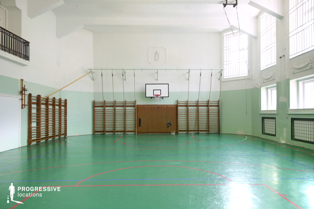This compact indoor gymnasium features a polished green linoleum floor with multiple court markings in red and blue, suited for varied school sports activities. The walls are finished in a pastel mint-green lower section with white upper areas, offering a soft and retro visual tone. Wooden wall bars line the walls, with hanging ropes and gymnastic rings suspended from metal beams, emphasizing its traditional school-sport function. Large, barred windows allow natural light to illuminate the space, while architectural moldings and an iron-wrought balcony add a subtle historical character. The central basketball hoop and padded wall suggest safety-conscious design, making this a functional and nostalgic location ideal for school-themed productions or retro-inspired visual projects.