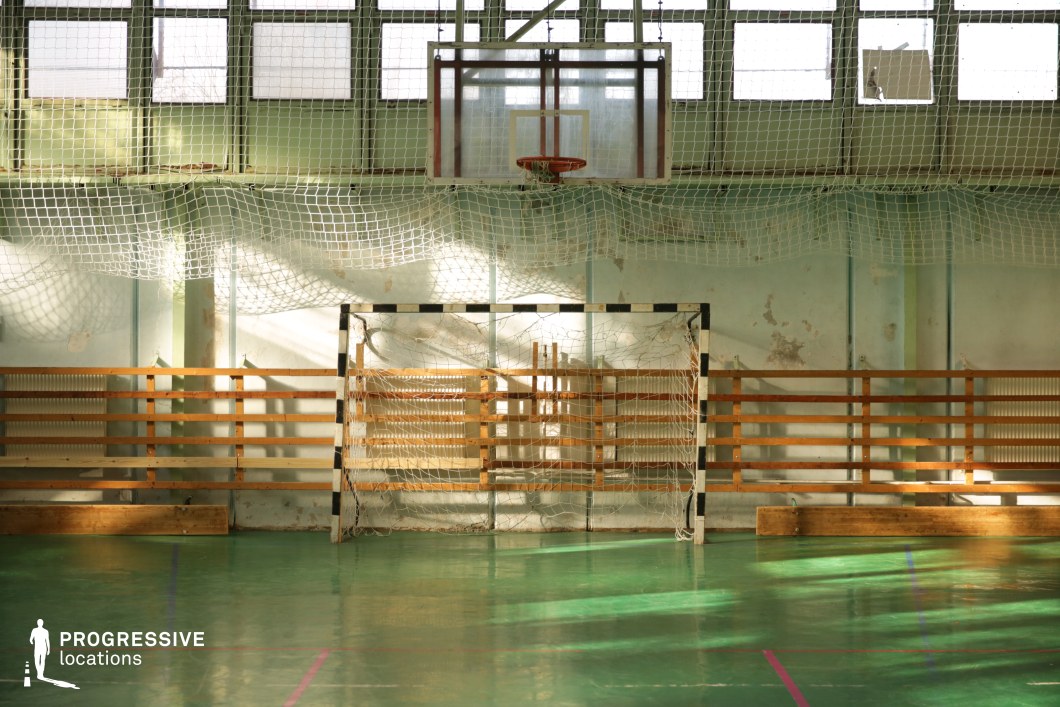 This indoor sports hall presents a timeworn but atmospheric setting, characterized by its glossy green flooring and visible signs of aging on the pale green walls. The focal point is a black-and-white goalpost placed directly beneath a suspended basketball backboard, both fitted with worn nets and protective netting extending along the wall. Wooden slatted barriers line the back wall, adding to the utilitarian, almost vintage look of the space. The room is softly lit by high-set frosted windows, allowing diffused natural light to cast dynamic shadows across the court. The subtle wear and imperfections on the surfaces contribute to an authentic, unpolished ambiance, making this location especially suitable for gritty, realistic sports scenes, nostalgic school settings, or character-driven productions seeking a textured, raw backdrop.