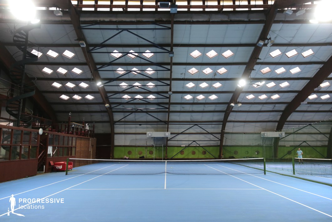 This image captures a front-facing view of an indoor tennis court hall characterized by its evenly arched, industrial-style roof supported by exposed steel trusses and wooden beams. The roof is punctuated by numerous rectangular skylights arranged in geometric patterns, enhancing the rhythm and symmetry of the space. The court surface is a uniform light blue, sharply demarcated with white lines, conveying a clean and professional aesthetic. The far wall and side panels exhibit a mix of green and metallic tones, contributing to an energetic yet functional environment. Ventilation units and industrial ducting are visibly integrated into the structure, reinforcing its utilitarian character. On the left side, a mezzanine with wood-framed glass enclosures and a spiral staircase adds architectural interest and offers elevated vantage points for observation or filming. The overall scale is generous and open, well-suited for dynamic camera angles or wide framing. The consistent geometry and lighting conditions make this space particularly versatile for sport-related shoots, promotional videos, or any creative project requiring a structured, indoor athletic setting.