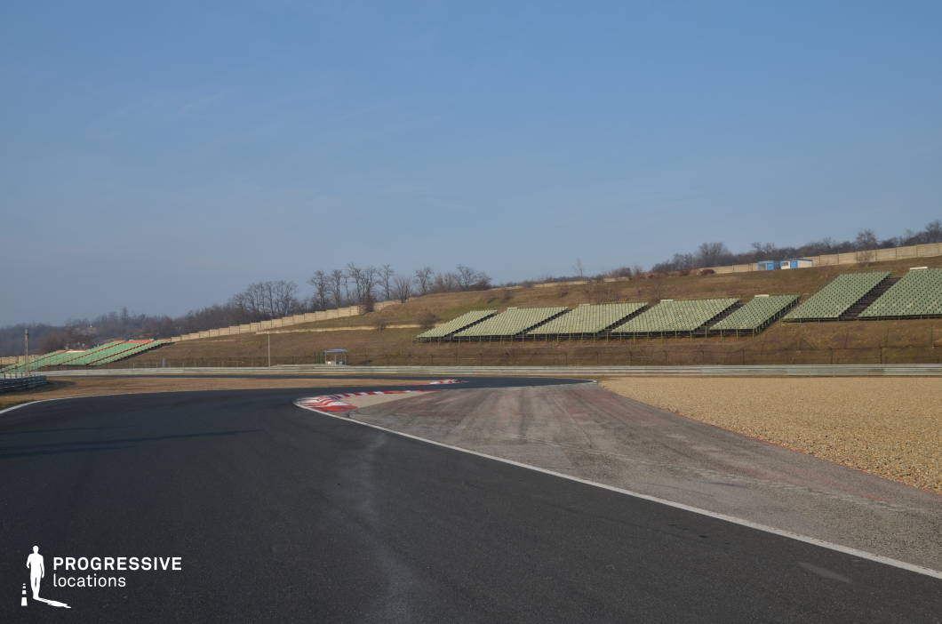 This location features a wide asphalt racetrack curving sharply, bordered by runoff areas with contrasting textures and colors. Tiered grandstands rise from the surrounding hillside, offering strong visual symmetry and a sense of scale, while the gentle slope and fencing define the space with clean lines. The openness and layout allow for dynamic movement and long-lens shots. This is an ideal filming location for sports dramas, action sequences, or car commercials, particularly those centered on racing, training montages, or high-speed chase scenes.