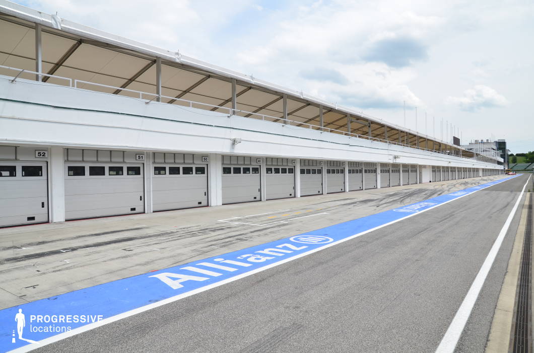 This expansive concrete pit lane stretches alongside a long row of garages, marked by vivid track lines and tire marks that hint at high-speed activity. Opposite, tiered grandstands rise under a series of repeating canopy structures, all separated by reinforced fencing and barriers. The composition offers strong linear depth and a sleek, technical atmosphere. This location is ideal for action films, sports documentaries, car commercials, or high-stakes racing scenes requiring an authentic and professional motorsport environment.
