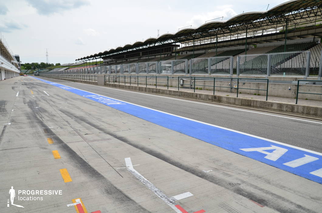 This expansive concrete pit lane stretches alongside a long row of garages, marked by vivid track lines and tire marks that hint at high-speed activity. Opposite, tiered grandstands rise under a series of repeating canopy structures, all separated by reinforced fencing and barriers. The composition offers strong linear depth and a sleek, technical atmosphere. This location is ideal for action films, sports documentaries, car commercials, or high-stakes racing scenes requiring an authentic and professional motorsport environment.