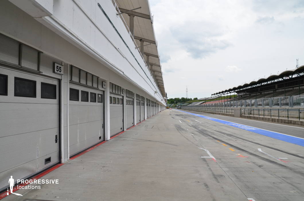 This long, symmetrical stretch of concrete pit lane is lined with uniform garage doors and overhangs, set opposite spectator stands and separated by fencing and a clearly marked blue pit lane. The expansive layout and clean, structured lines emphasize speed, precision, and performance. This location is ideal for filming contemporary racing scenes, sports documentaries, high-stakes action sequences, or automotive commercials, especially those involving pit stop choreography, technical preparations, or behind-the-scenes racing drama.