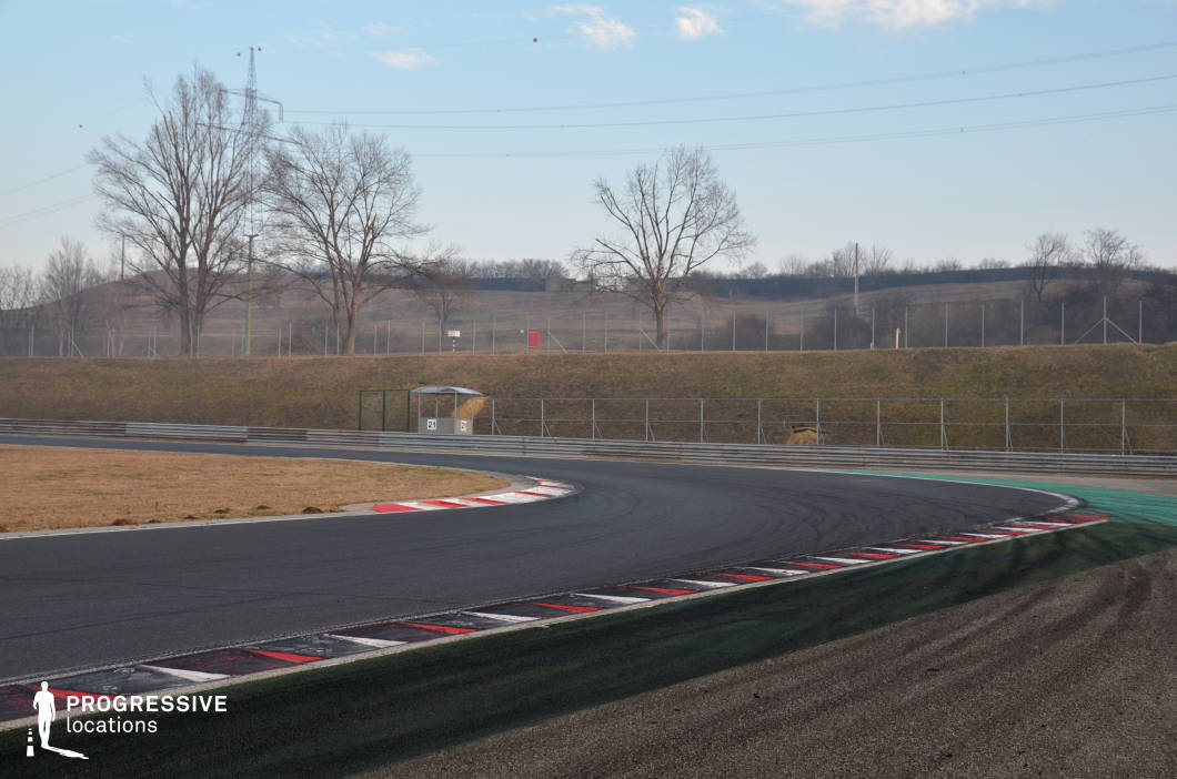 This image captures a dynamic curve on a modern racing circuit, complete with precision-painted curbs, layered tire marks, and safety barriers enclosing the track. The surroundings are open and slightly elevated, offering wide visibility and a sense of motion even in stillness. The bend’s technical design and clean infrastructure make it ideal for capturing high-speed maneuvers, dramatic overtakes, or chase sequences. This location suits action films, racing-centric narratives, or promotional content focusing on automotive performance, precision, and adrenaline.