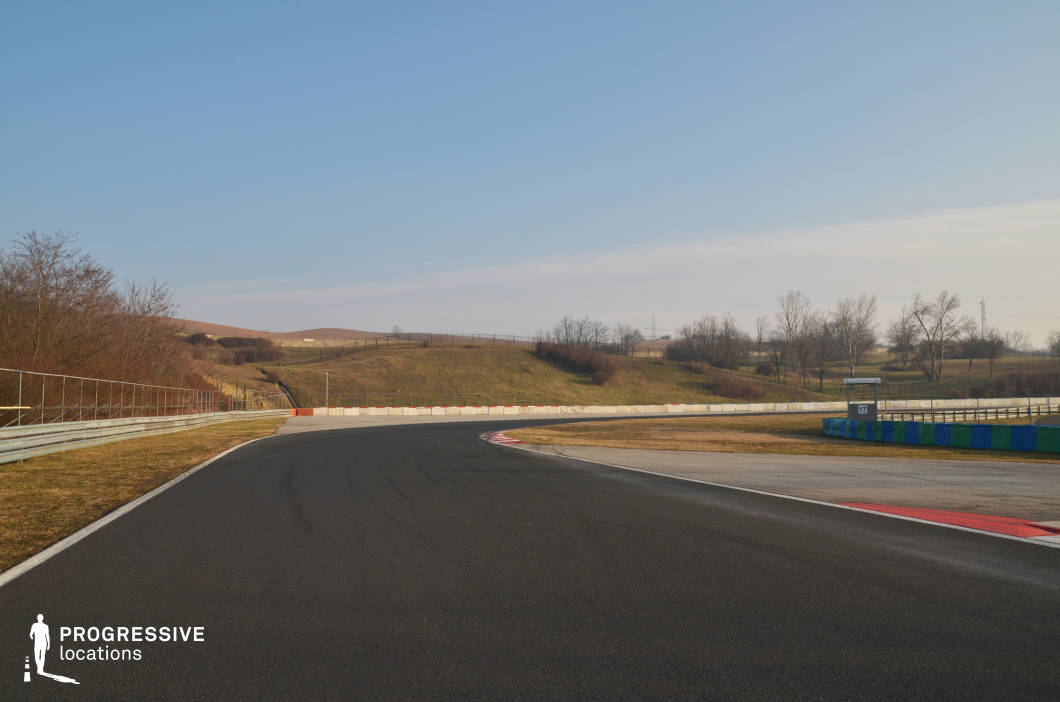 A wide, smooth curve of asphalt winds through gently rolling terrain, flanked by safety barriers and runoff areas. The open landscape enhances visibility and scale, while subtle track markings and tire streaks suggest high-speed action. The clean, expansive layout is ideal for dynamic driving shots, drone footage, or wide-angle racing sequences. This location suits car commercials, racing films, or action scenes requiring long tracking shots and controlled high-speed movement in a visually open environment.