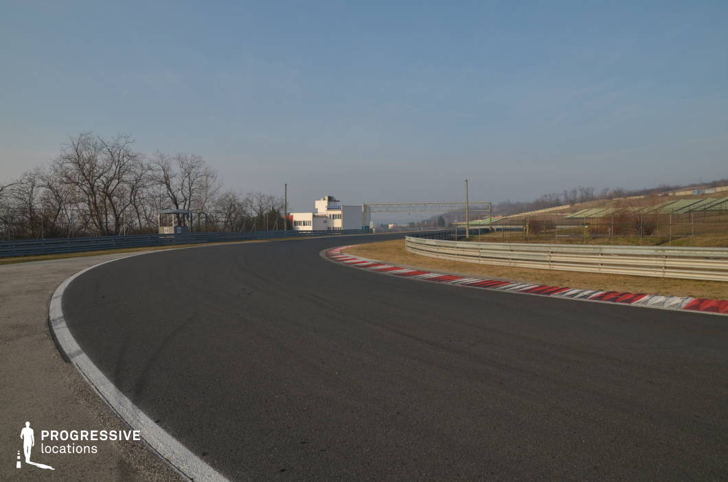 This location highlights a wide, sweeping bend on a professional racetrack, bordered by striped curbs, safety barriers, and open embankments. The unobstructed view toward the horizon and the layered terrain offer a sense of speed and motion, enhanced by the clean tarmac and precise track markings. Its spaciousness and visual clarity make it well-suited for filming dynamic driving sequences, test runs, or car commercials, particularly for genres such as action, motorsports dramas, or high-octane thrillers.
