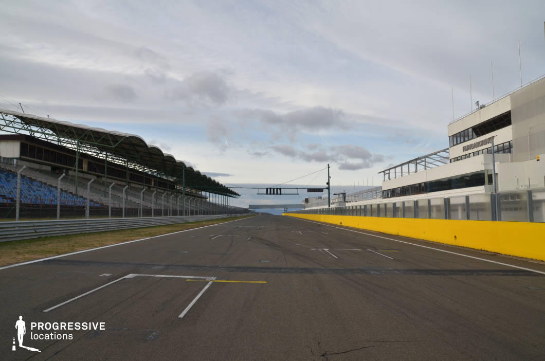This wide-angle image showcases a modern motorsport racetrack captured from the start-finish line, flanked by grandstands on one side and a multi-level control building on the other. The asphalt surface is marked with precise white and yellow racing lines, while safety barriers and tall fencing enhance the sense of a high-speed, regulated environment. The architecture on both sides is sleek and functional, with the left side featuring a curved steel canopy over rows of blue seating, and the right side dominated by white modular structures. The deep vanishing point and symmetrical composition create a dynamic sense of depth and speed. This location is ideal for filming automotive content, action sequences, or competitive sports dramas, offering a visually striking and versatile backdrop.