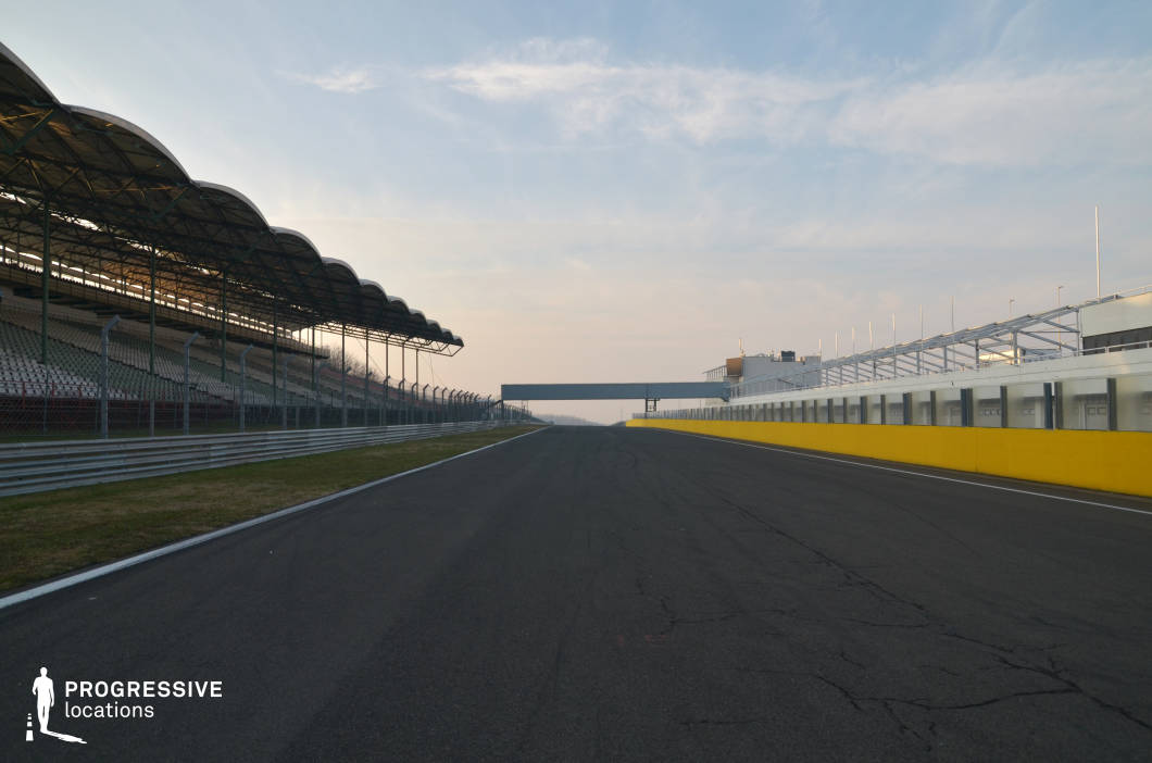 This expansive asphalt track is flanked by grandstands on one side and a long row of pit garages on the other, clearly defining the start-finish straight of a professional racing circuit. The structural elements—metal fencing, overhead signage, and modernist canopies—convey a highly technical, competitive environment. The scale and perspective emphasize speed and anticipation, making it an ideal filming location for action-packed racing scenes, sports dramas, futuristic car sequences, or even high-stakes escape scenarios in thrillers or heist films.