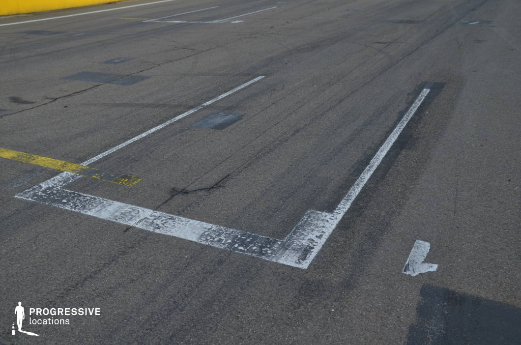 This image captures a close-up view of a motor racing circuit's start grid, with clearly defined white and yellow painted lines over worn asphalt, marked by tire streaks and subtle surface cracks. The texture and wear of the pavement, along with the repetitive grid layout, convey speed, precision, and tension. The setting provides an ideal backdrop for racing sequences, sports dramas, or commercial shoots involving motorsports, with potential for dramatic start-line moments, slow-motion takeoffs, or intense close-up action.