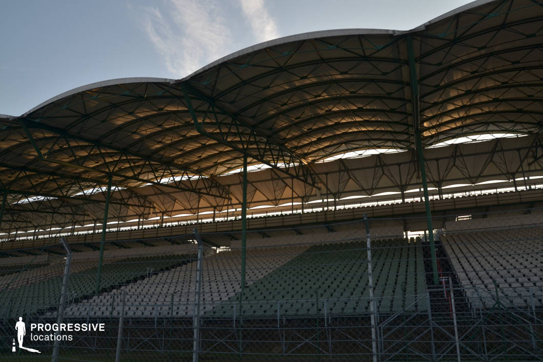 This location showcases a large, modern grandstand featuring a sweeping wave-patterned canopy supported by a web of green steel beams. The repeating rhythm of the seating rows and the semi-transparent roof create a dynamic interplay of light and shadow. The scale and architectural design suggest a professional sporting venue, adding visual energy and contemporary realism. Ideal for filming high-stakes sports dramas, racing sequences, concert scenes, or dystopian thrillers, this setting offers a dramatic backdrop for both large-scale events and emotionally charged moments in the stands.