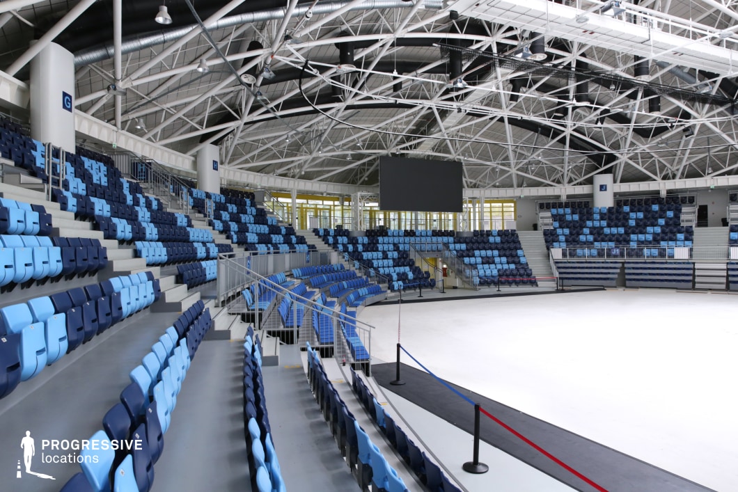 This photo captures a detailed side view of a modern indoor arena, emphasizing its structured layout and architectural clarity. The seating is arranged in tiered rows and features a dual-tone color scheme of navy blue and light blue, creating a visually appealing pattern. Overhead, the intricate lattice of white steel beams and exposed ducts forms an industrial-style ceiling, equipped with spotlights and a suspended central scoreboard. The arena floor is currently uncovered, providing a blank canvas suitable for a wide range of event setups. The space is optimal for multi-purpose indoor shoots or productions requiring symmetrical design, clear sightlines, and a clean, adaptable setting.
