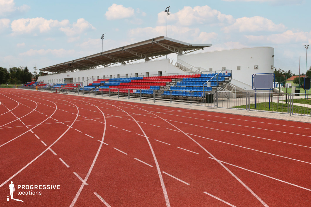 This image features a small-scale outdoor athletics stadium, centered around a bright red synthetic running track with clearly marked white lanes. Adjacent to the track, aluminum bleachers provide tiered seating with a bold pattern of blue and red plastic chairs. A low, modern white structure behind the stands offers basic facilities and is topped with a cantilevered metal roof for partial shade. The overall design is clean and functional, suitable for local sports events, training sessions, or youth competitions. Its compact size and organized structure make it a versatile location for shoots requiring a simplified, accessible stadium backdrop.