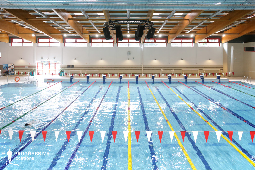 This image presents a modern indoor swimming pool facility with a balanced combination of functionality and visual clarity. The main focus is a standard-length competition pool, divided into multiple lanes by vibrant red, blue, yellow, and green lane dividers, clearly marking the structure’s use for regulated swimming events. The view is symmetrical and centered, emphasizing the pool’s geometric order and proportionality. The surrounding architecture features white walls and large red-framed windows positioned high along the perimeter, allowing diffused natural light to enter. A light timber grid ceiling with exposed ductwork and mounted lighting adds to the venue’s technical and clean appearance. The acoustic ceiling design includes a black circular speaker array, suspended above the water for optimal sound projection. Bleacher-style wooden benches line the back wall, offering practical seating with a minimal visual footprint. The material palette of wood, ceramic tiles, metal fittings, and glass contributes to a modern, efficient atmosphere without visual clutter. This space is ideal for productions in need of a bright, contemporary aquatic environment with symmetrical composition and clear structural elements. The clean layout and well-lit conditions provide versatile conditions for both static and motion-oriented shoots.