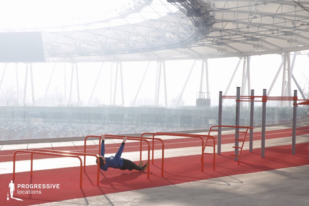 This visually dynamic image showcases a modern outdoor calisthenics and functional fitness area within a stadium complex. The red rubberized flooring defines a designated workout strip, equipped with steel bars in orange and grey, suited for exercises like pull-ups, dips, or bodyweight rows—as demonstrated by a lone athlete mid-rep in the foreground. The equipment is neatly arranged for circuit-style movement and spatial flow. The surrounding stadium architecture features tall, white tensile supports and a transparent glass railing, maintaining visibility while offering a sense of enclosure. Above, a canopy structure filters light across the scene, while soft atmospheric haze in the distance mutes the large video board and stadium seating, allowing the training space to remain the visual focal point. This location is ideal for productions centered on athleticism, performance, or individual training. The contrast between the bold color palette, structural repetition, and soft natural light offers a visually rich, contemporary backdrop for high-energy or lifestyle-focused content.