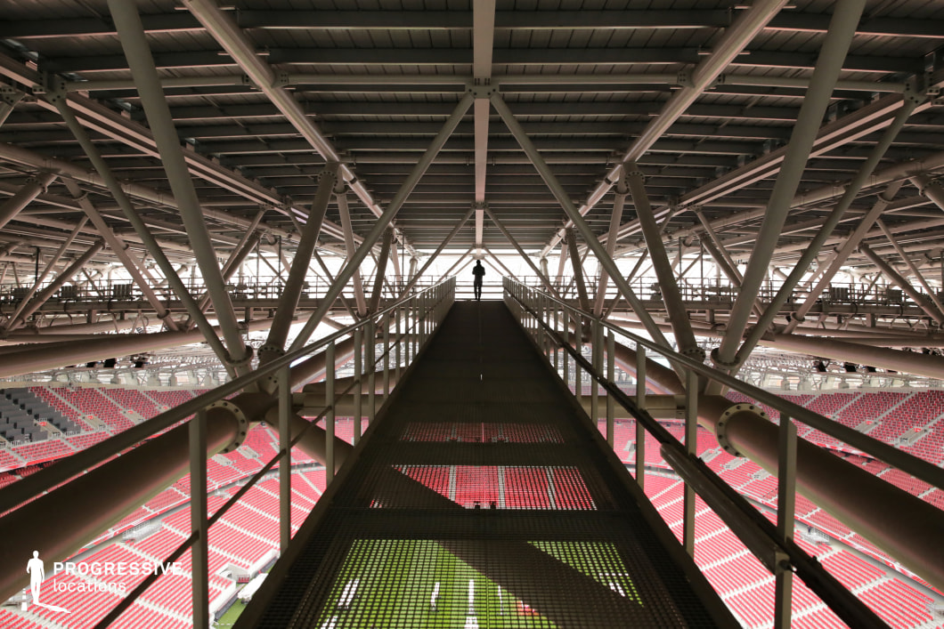 This image offers a rare perspective from the upper catwalk of a large stadium, emphasizing the complex network of steel trusses and architectural beams that support the structure's roof. A suspended metal walkway leads toward the center of the frame, creating a strong sense of depth and symmetry, with a single silhouetted figure at the vanishing point enhancing the spatial drama. The overhead industrial lighting and mechanical systems are exposed, highlighting the raw engineering aesthetics. Beneath the walkway, partially visible through the gridded metal floor, the red spectator seats and the green pitch of the stadium hint at the scale and function of the venue. This unique viewpoint is ideal for productions focused on architectural engineering, behind-the-scenes infrastructure, or dramatic, introspective compositions that play with height and structural repetition. The scene balances monumental industrial design with a cinematic, almost sci-fi quality.