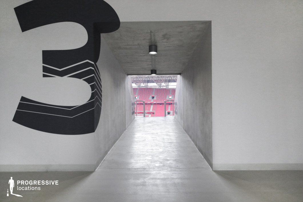 This photo captures the clean, minimalist interior of a stadium tunnel leading to the spectator area, emphasizing geometric simplicity and modern architectural lines. The corridor features smooth concrete flooring and walls, illuminated by simple cylindrical ceiling lights. A bold, stylized number 