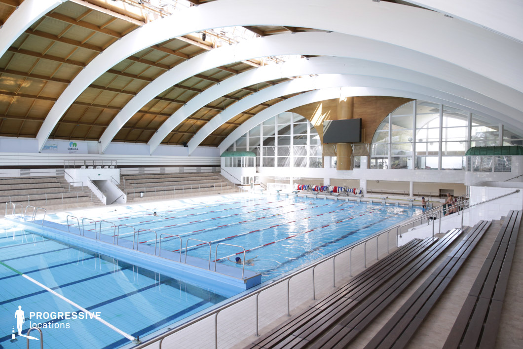 This indoor swimming facility is defined by its striking architectural design, featuring large white arched beams that span the entire width of the hall, complemented by warm wooden ceiling panels. The pool area includes multiple swim lanes with marked dividers and depth indicators, making it suitable for professional aquatic activities such as swim training, competitions, or synchronized swimming. The spectator stands are constructed with concrete terraces and simple wooden bench seating, offering a clean, utilitarian look. Large arched windows and glass walls on the far end allow ample natural light to filter in, giving the space an airy and expansive atmosphere. Functional elements like safety rails, storage for lane buoys, and a digital scoreboard contribute to the location’s authenticity as a competitive sports environment. Versatile and architecturally distinct, this venue is ideal for dynamic water-based filming or athletic-themed commercial productions.