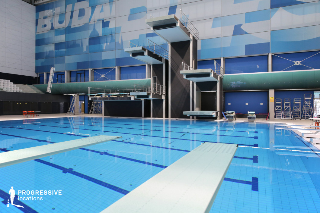 This indoor aquatic facility features a state-of-the-art diving platform complex set within a spacious and architecturally unified environment. The main focus is a deep blue-tiled diving pool surrounded by various levels of springboards and platforms, ranging from low to high elevations, designed for professional diving competitions. The image captures the scene from a low corner angle, enhancing the vertical emphasis of the towering structures and their reflections in the still water. The backdrop is a large-scale wall mural composed of abstract blue gradients and stylized architectural silhouettes, adding a dynamic visual identity to the space. The materials include non-slip composite on the boards, reinforced concrete supports, and brushed metal railings, all contributing to a robust, performance-oriented aesthetic. Surrounding elements such as tiered spectator seating, movable ladders, and technical equipment remain discreetly arranged along the perimeter, allowing the central diving area to remain visually dominant. The overall color palette is a mix of blues, greys, and whites, reinforcing the facility’s modern and aquatic atmosphere. This venue is ideal for productions that require a visually striking, technically precise location with a strong vertical composition and professional sports infrastructure.