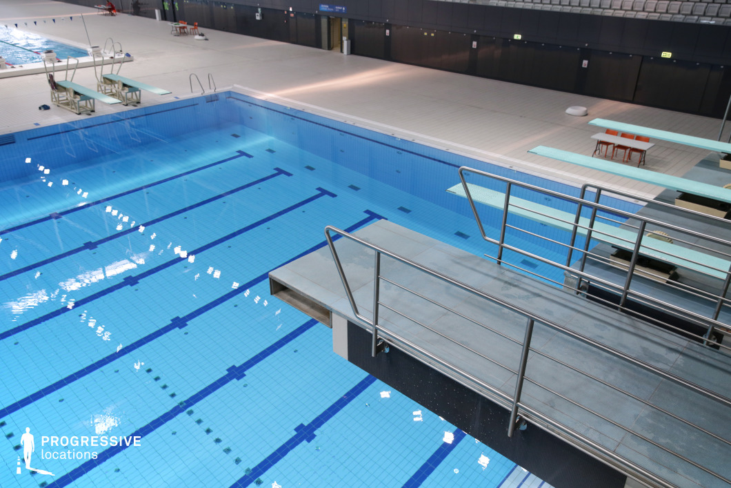 This indoor facility features a diving and training pool setup within a modern aquatic sports complex. The image showcases multiple diving boards at varying heights, with a central focus on a raised platform constructed from slip-resistant material and framed with stainless steel railings. The geometric layout of the pool, complete with deep blue lines and clearly marked zones, supports technical training and regulated diving practice. Surrounding the pool is a wide, tiled deck area providing ample operational space. The subdued color palette—light blues, greys, and whites—contributes to a clean, focused environment. The upper spectator seating area is subtly visible in the background, while the lighting is evenly distributed through ceiling-mounted fixtures, ensuring optimal visibility without visual distraction. The photo is taken from a slightly elevated diagonal angle, offering a clear perspective on the structural hierarchy of the boards and the water’s reflective quality. The design prioritizes functionality, symmetry, and precision, ideal for high-performance sports environments. This venue is well-suited for visual content requiring an authentic, high-end aquatic training backdrop with a structured, minimal aesthetic.