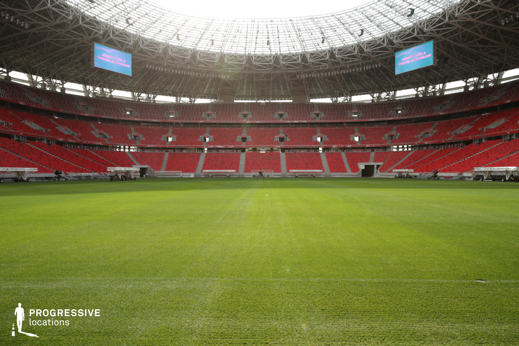 Captured from field level, this image presents a centered, wide-angle view of a contemporary football stadium’s interior. The expansive grass pitch stretches into the foreground, meticulously maintained with subtle mowing lines that add texture and directionality. Encircling the field are striking red seating tiers, divided across multiple levels, with the architecture emphasizing symmetry and scale. The steel roof trusses span the open-air ceiling, supporting large-format LED displays that hover above the spectators' zone. The layout is balanced by two main player tunnels positioned opposite each other, directly aligned and enhancing the axial composition. The lighting is natural, with daylight streaming through the roof structure and subtly illuminating the field and seats. This setup is ideal for productions requiring grand sports arena visuals or symbolic wide-open spaces. The clean lines, strong geometry, and monumental atmosphere make it especially effective for establishing shots, promotional material, or content emphasizing performance and scale.