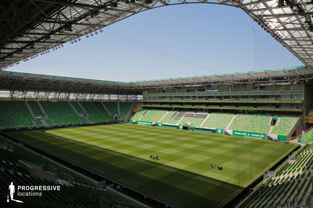 This football stadium presents a modern, structured environment featuring a full bowl of vibrant green seating arranged in layered tiers. A transparent roof canopy supported by a steel frame spans the perimeter, allowing natural light to flood the space while ensuring coverage. The opposing main stand houses premium boxes and media facilities, enhancing its visual and functional prominence. The field is meticulously maintained, with alternating striped grass patterns adding visual texture. The uniform color palette of greens and grays creates a clean, cohesive look suitable for both sporting event shoots and commercial productions. Its scale, symmetry, and architectural openness make it ideal for large-scale cinematic visuals and broadcast-style compositions.