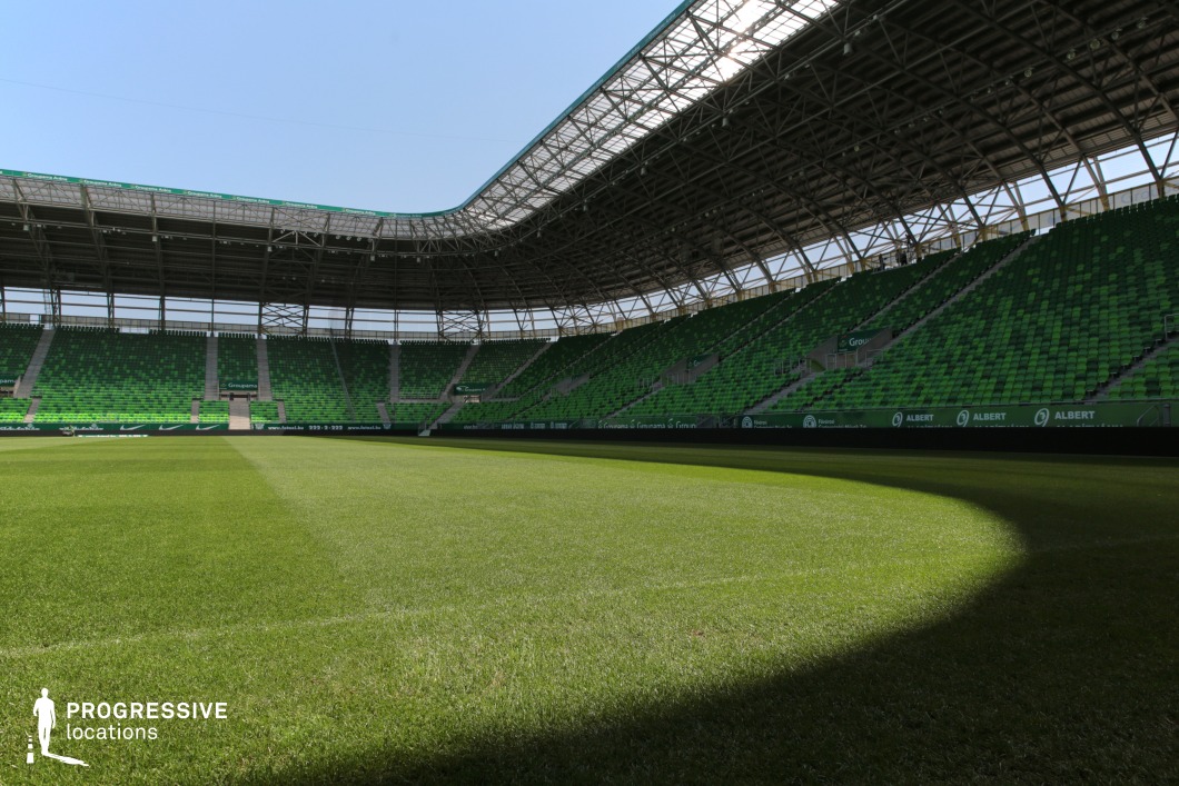This image presents a medium-to-large football stadium characterized by its modern architectural features and uniform visual style. The interior view, captured from pitch level, emphasizes the curvature of the seating arrangement and the partially enclosed roof structure. The spectator stands are clad in a vibrant gradient of green seating, arranged in a pixel-like pattern that provides both visual depth and brand consistency. The stadium’s defining structural element is its sweeping, curved steel canopy supported by an exposed truss system, which offers partial coverage and introduces dramatic, rhythmic lines to the composition. The transparent roof sections contribute to a light, open-air feel, enhancing visibility while maintaining a sense of enclosure. The camera angle, taken low and slightly angled from the field, foregrounds the quality of the natural grass surface and accentuates the scale and slope of the surrounding stands. The absence of central elements or field markings ensures a clean visual field ideal for compositional flexibility. This location is especially fitting for productions requiring a bold, cohesive backdrop with a dynamic interplay of color, structure, and spatial openness, particularly within a sports-themed or architecturally striking context.