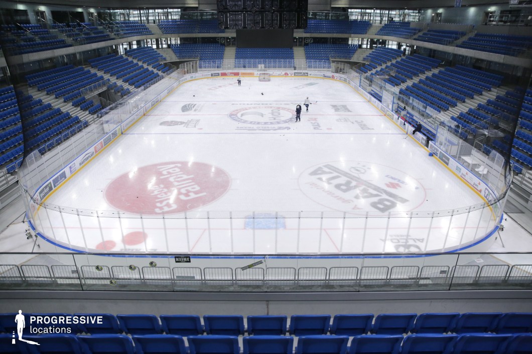 The image showcases a contemporary indoor ice hockey arena with a full-sized rink enclosed by transparent safety boards and netting. The stands feature symmetrical arrangements of blue plastic seats on both sides, creating a bold visual rhythm against the white ice surface. The rink displays various center-ice advertisements and regulation markings, framed by yellow kickplates and plexiglass barriers. Overhead, a large central scoreboard hangs above the rink, and the arena is well-lit by integrated ceiling fixtures. This location offers a highly professional setting ideal for shooting sports sequences, commercial spots, or scenes requiring a sleek and structured athletic environment.