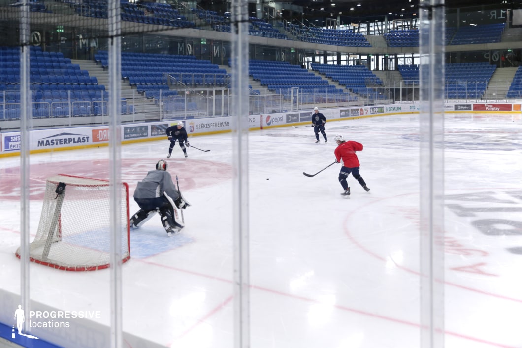 This image captures a close-up view of an ice hockey rink during a training session, featuring players in action, including a goaltender and three skaters engaged in a drill. The arena is enclosed with transparent plexiglass barriers and surrounded by symmetrical blue-tiered seating. The ice surface is professionally marked and well-maintained, with branding and advertisements displayed along the boards. The facility's modern structure includes concrete staircases, glass railings, and a high ceiling equipped with lighting rigs. The space is ideal for filming dynamic sports scenes, athlete-focused campaigns, or productions needing a high-energy, contemporary sports setting.