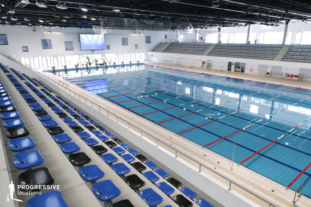 This image showcases a spacious indoor swimming complex featuring a full-size Olympic pool with marked lanes, surrounded by tiled decking and spectator seating. The pool's pristine turquoise water is sectioned by red and blue lane dividers, reflecting the clean geometry of the interior. Tiered spectator stands in blue and black seat tones extend along the poolside, offering clear sightlines for viewers. Above, the dark ceiling structure with exposed mechanical systems adds an industrial character, contrasted by abundant daylight pouring through large, upper-level windows and glass doors. A digital wall clock and scoreboard dominate the far wall, enhancing the venue’s functionality for competitions and large events. This versatile location is ideal for filming sports scenes, aquatic-themed visuals, or capturing clean architectural compositions with a bright, controlled lighting environment.