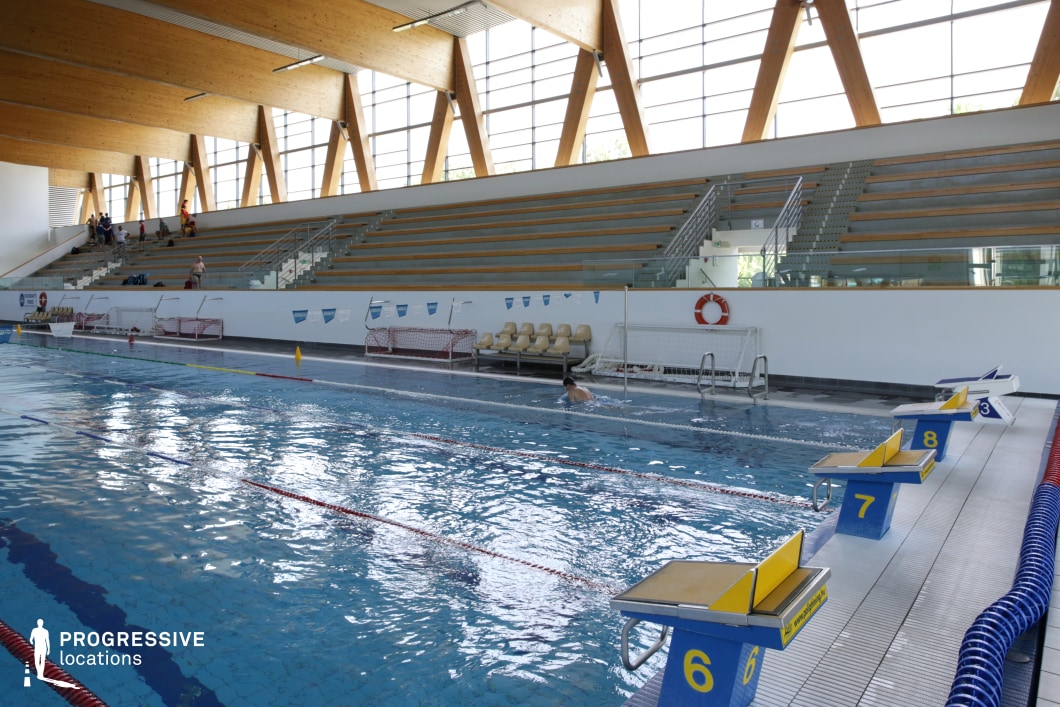 This indoor swimming facility features a competition-style pool, complete with starting blocks and lane dividers, making it suitable for aquatic sports and training scenes. The architecture is characterized by large, angled wooden trusses that support the roof and add warmth to the otherwise minimalistic space. Floor-to-ceiling windows along the upper perimeter flood the pool area with natural light while maintaining a clean, modern look. The grandstand seating is built with concrete and wooden benches in a stepped arrangement, offering clear sightlines across the pool. A modest section of molded spectator chairs provides additional versatility. Details like the wall-mounted lifebuoy, water polo goals, and practical stair access contribute to the functional authenticity of the setting. This location is ideal for shooting structured, athletic, or institutional scenes in a visually distinctive aquatic environment.
