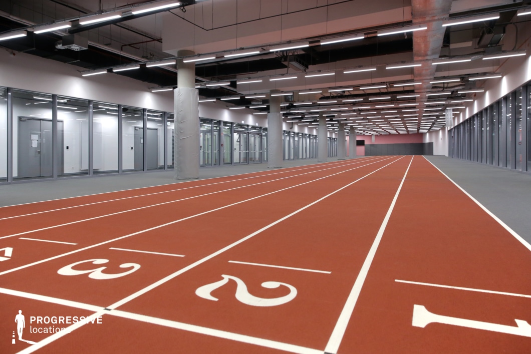 This image showcases a purpose-built indoor sprint track set within a high-tech training or athletic support facility. The red synthetic surface is divided into clearly marked lanes, each numbered and bordered with crisp white lines. The setting is defined by its modern, utilitarian architecture, featuring floor-to-ceiling glass partitions on both sides, which likely enclose administrative, medical, or team preparation areas. The ceiling is densely fitted with strip lighting and exposed infrastructure, including ductwork and piping, contributing to an industrial-tech aesthetic. Padded structural columns are spaced at regular intervals along the track’s edge, ensuring both spatial rhythm and safety. The layout is entirely linear, directing focus toward the vanishing point, which enhances the sense of acceleration and motion. This controlled, enclosed environment is ideal for performance testing, rehabilitation, or elite athlete warm-ups. Visually, it provides a clean, symmetrical backdrop perfect for stylized indoor sports content, dynamic movement sequences, or commercial projects requiring a sleek, high-performance atmosphere.